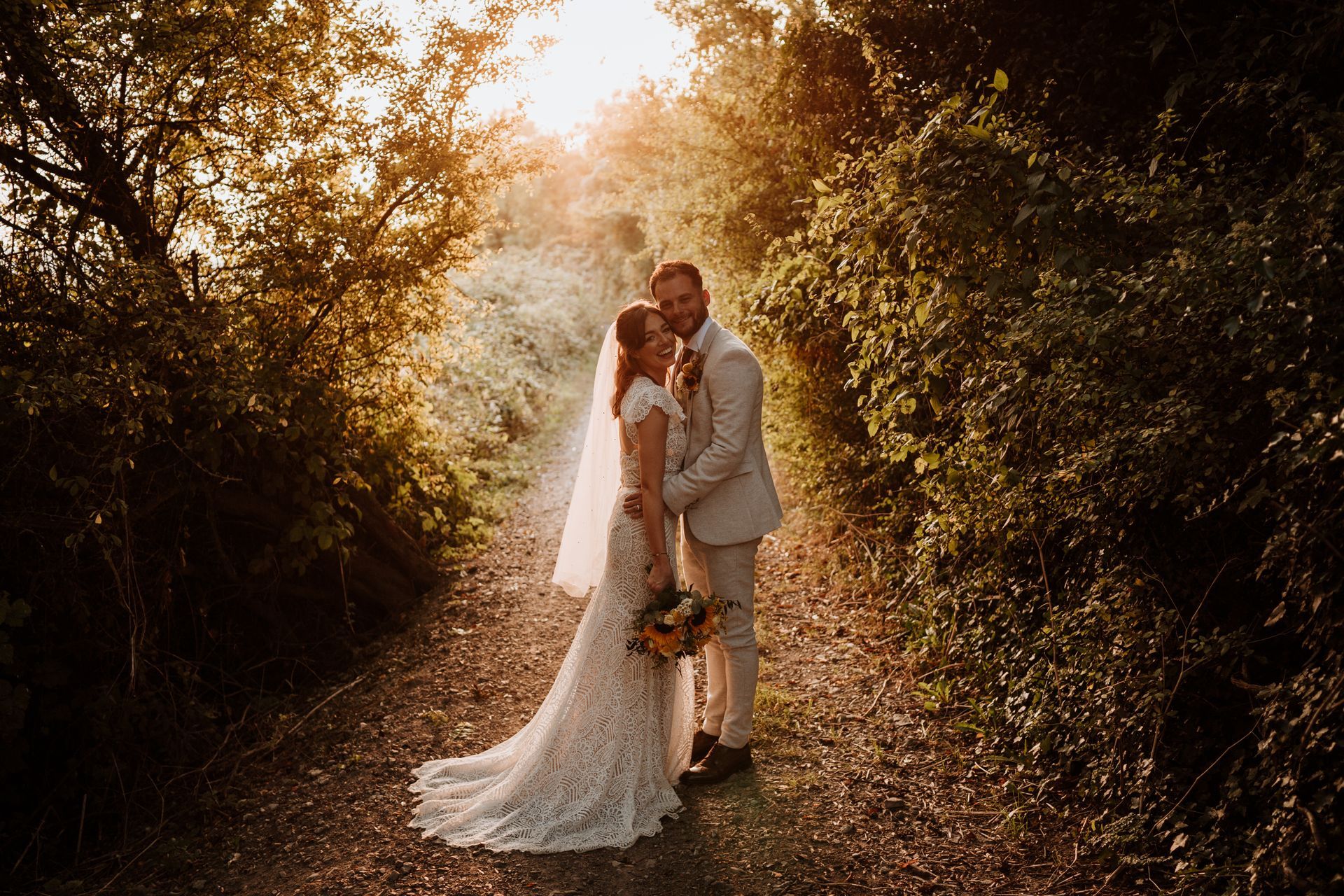 Bride and groom embracing on a sunlit forest path, surrounded by warm golden foliage