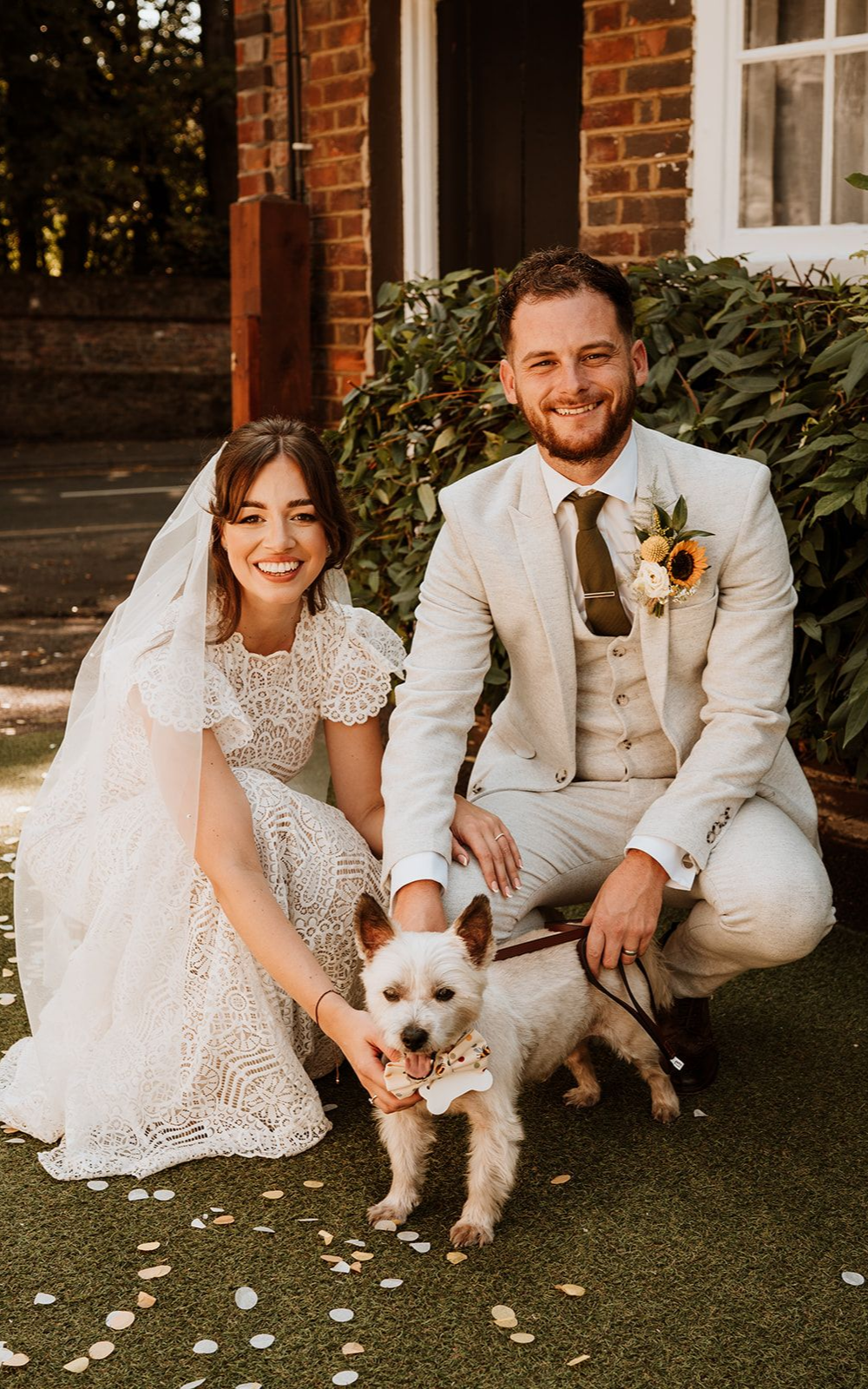 Bride and groom kneel outdoors with a small dog on a lawn in front of a brick house