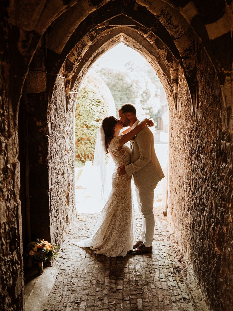 Couple embracing in a sunlit stone archway, with the bride in a lace gown and veil.