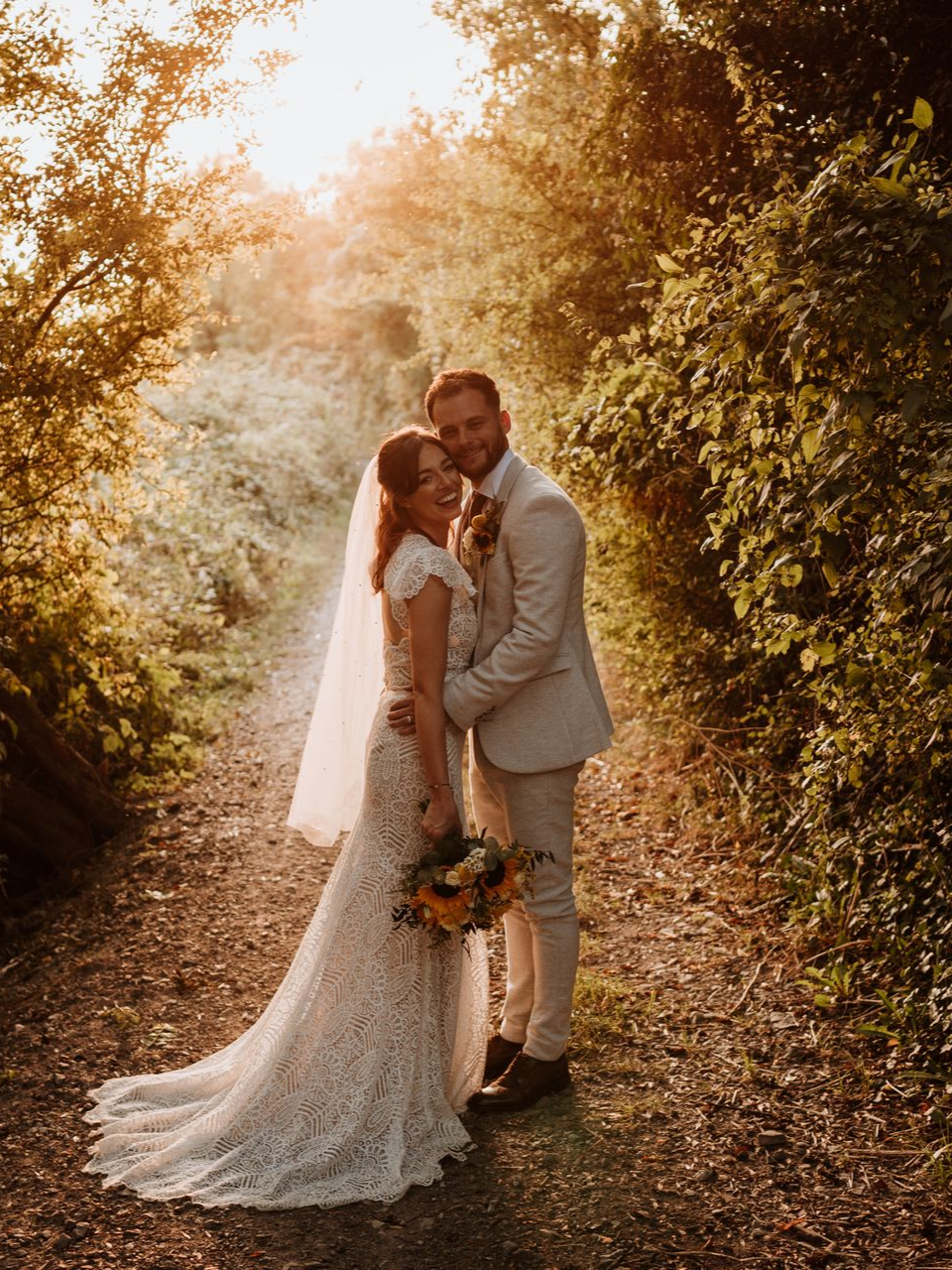 Bride and groom kissing on a sunlit forest path, with a long veil and bouquet.