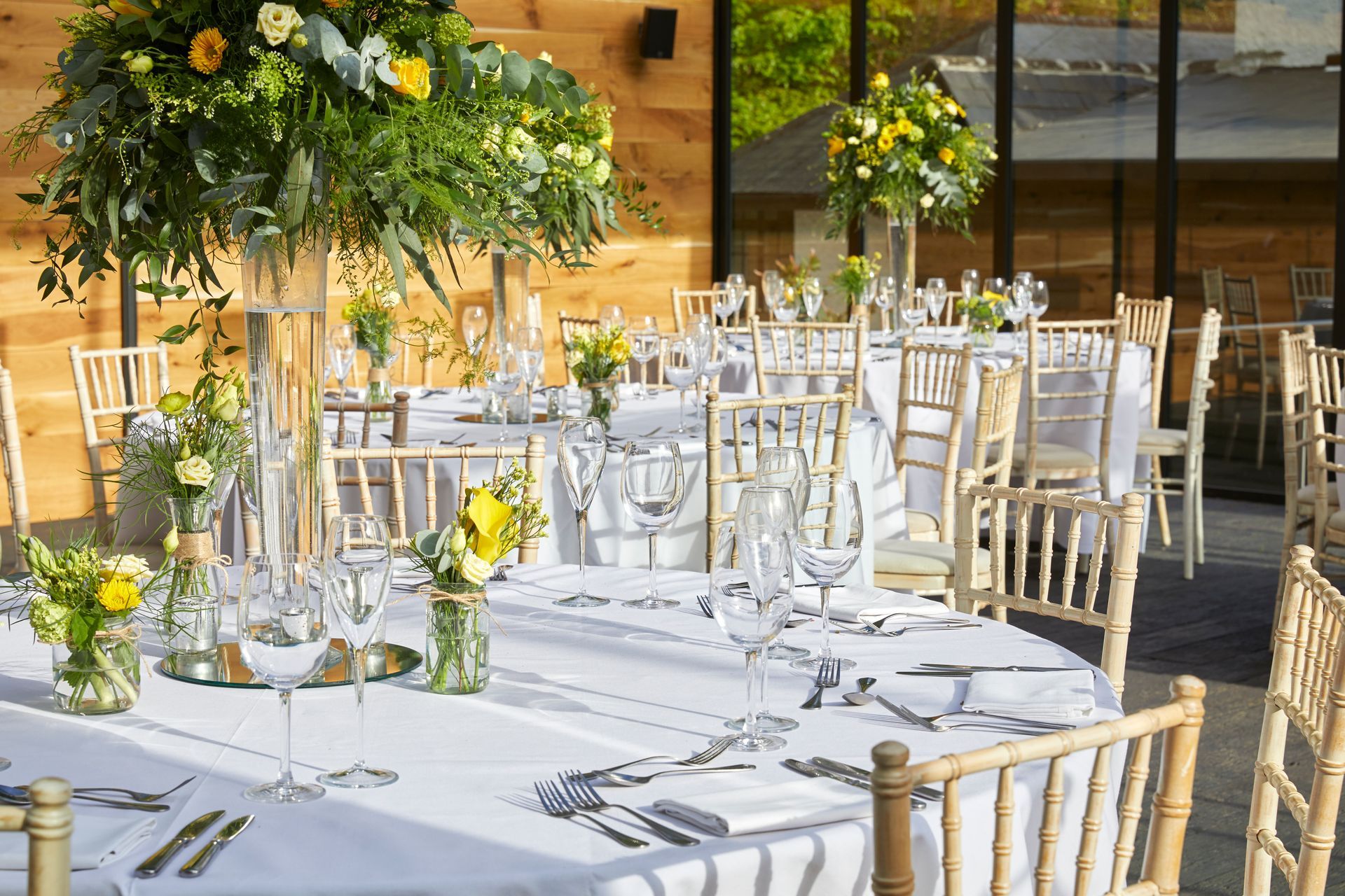 Outdoor wedding reception tables with white chairs, glassware, and yellow floral centerpieces in warm sunlight