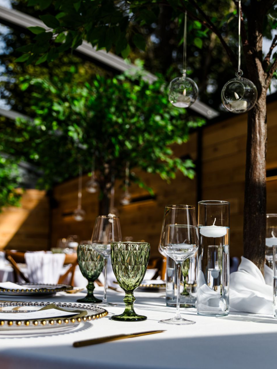 Elegant outdoor dining table with white tablecloth, glassware, plates, and green foliage overhead
