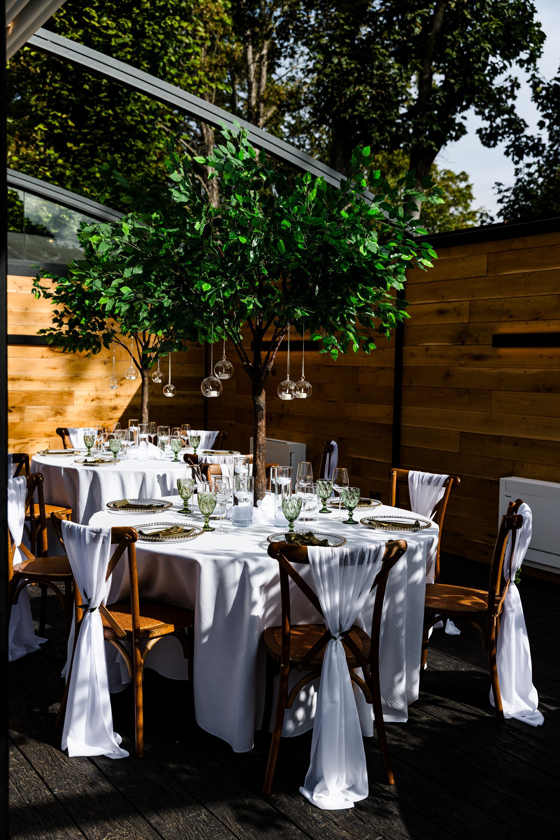 Round outdoor dining table set under a leafy tree, with white linens, brown chairs, and a wooden fence.