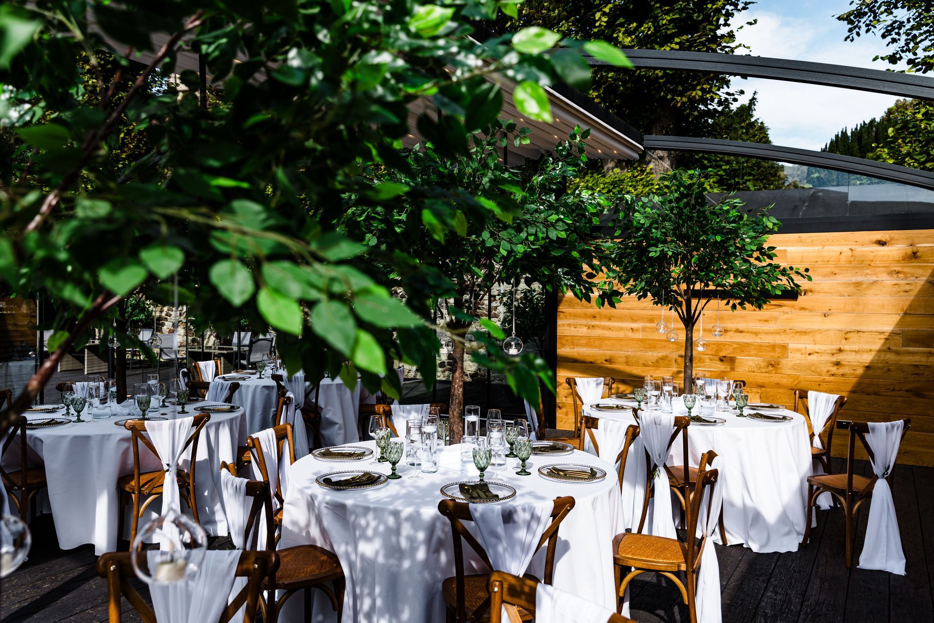 Outdoor dining area with round white-clothed tables, wooden chairs, and leafy greenery overhead