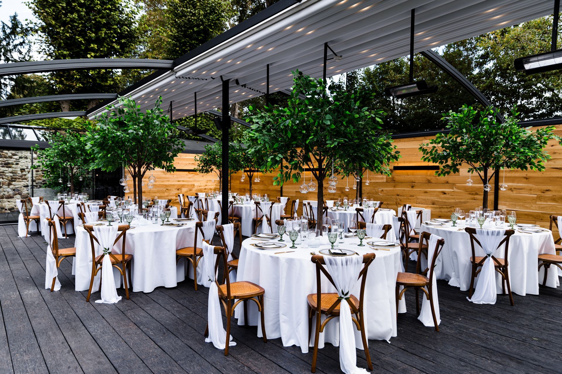 Outdoor event dining area with white-clothed tables, wooden chairs, and string lights under a covered patio