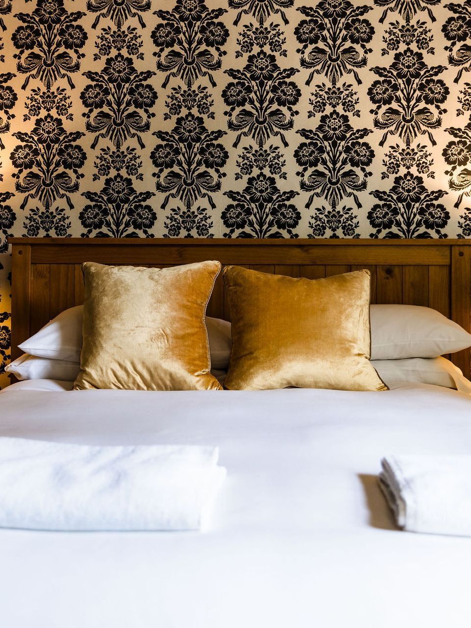 Neatly made bed with white linens, gold pillows, and ornate black wallpaper behind the headboard