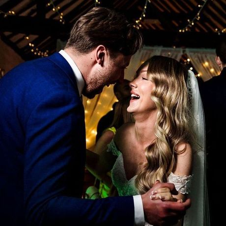 2026 wedding packages Couple dancing at wedding, man kisses woman's forehead, she smiles. Dimly lit barn setting, string lights in background.