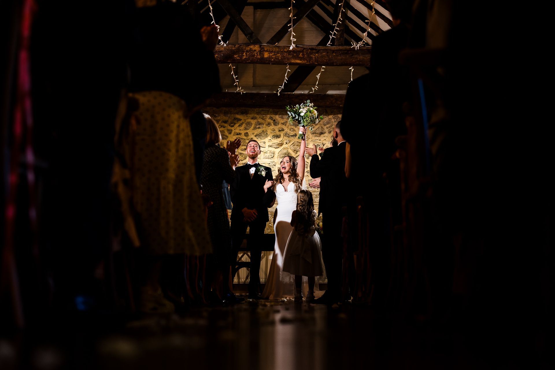 People in formalwear under a decorated archway at a dimly lit indoor wedding venue