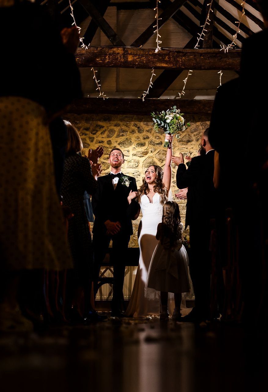 Wedding couple celebrating under rustic wooden beams, bride raising bouquet in a dimly lit barn aisle.