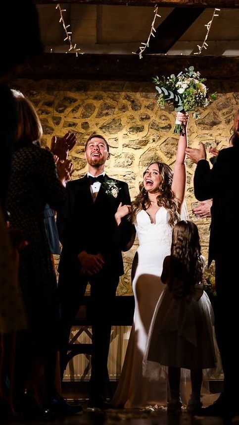 Newlyweds celebrate at their reception, raising their arms with guests applauding in a stone-walled venue