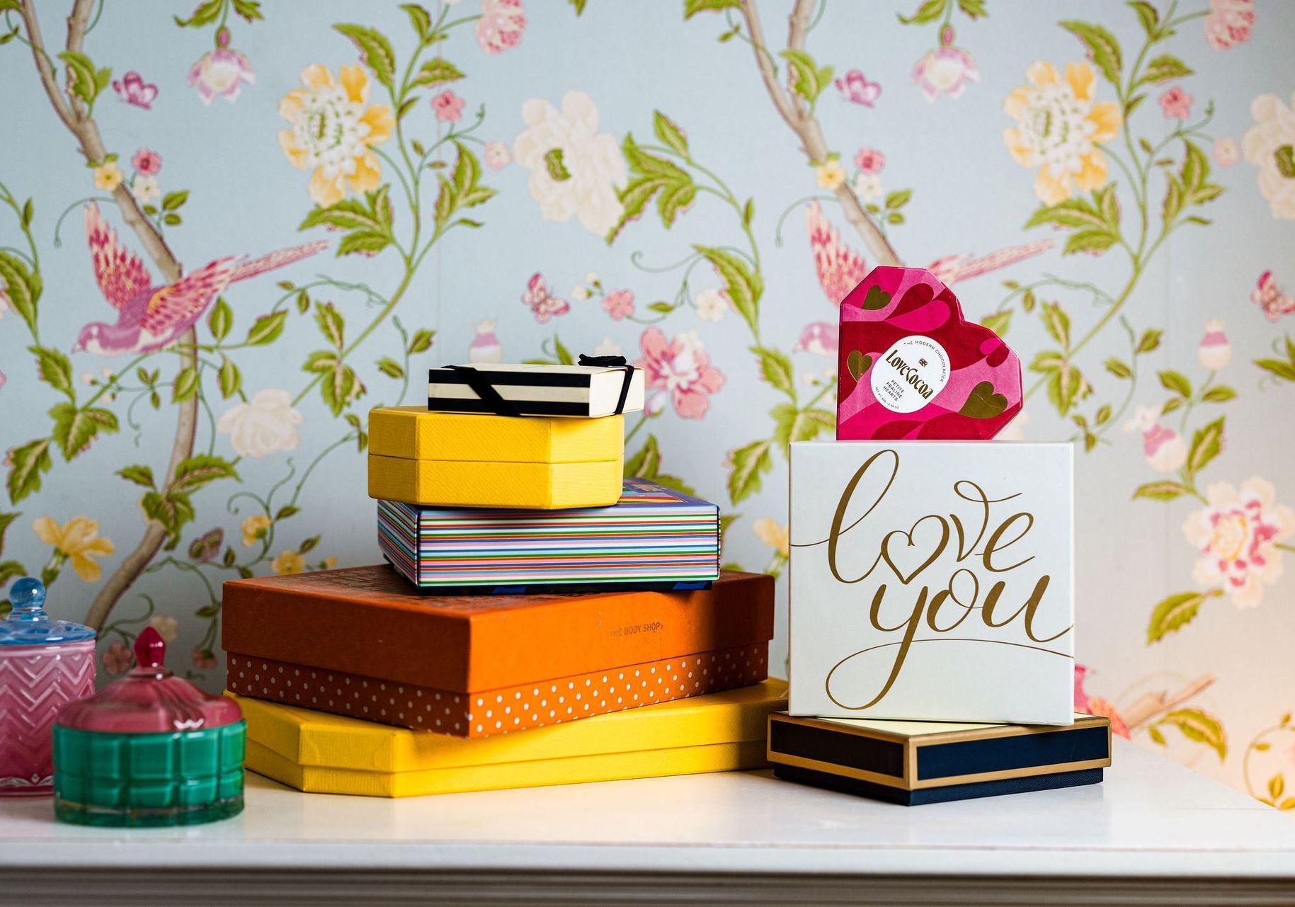 Colorful stacked gift boxes and a “love you” card on a white shelf against floral wallpaper