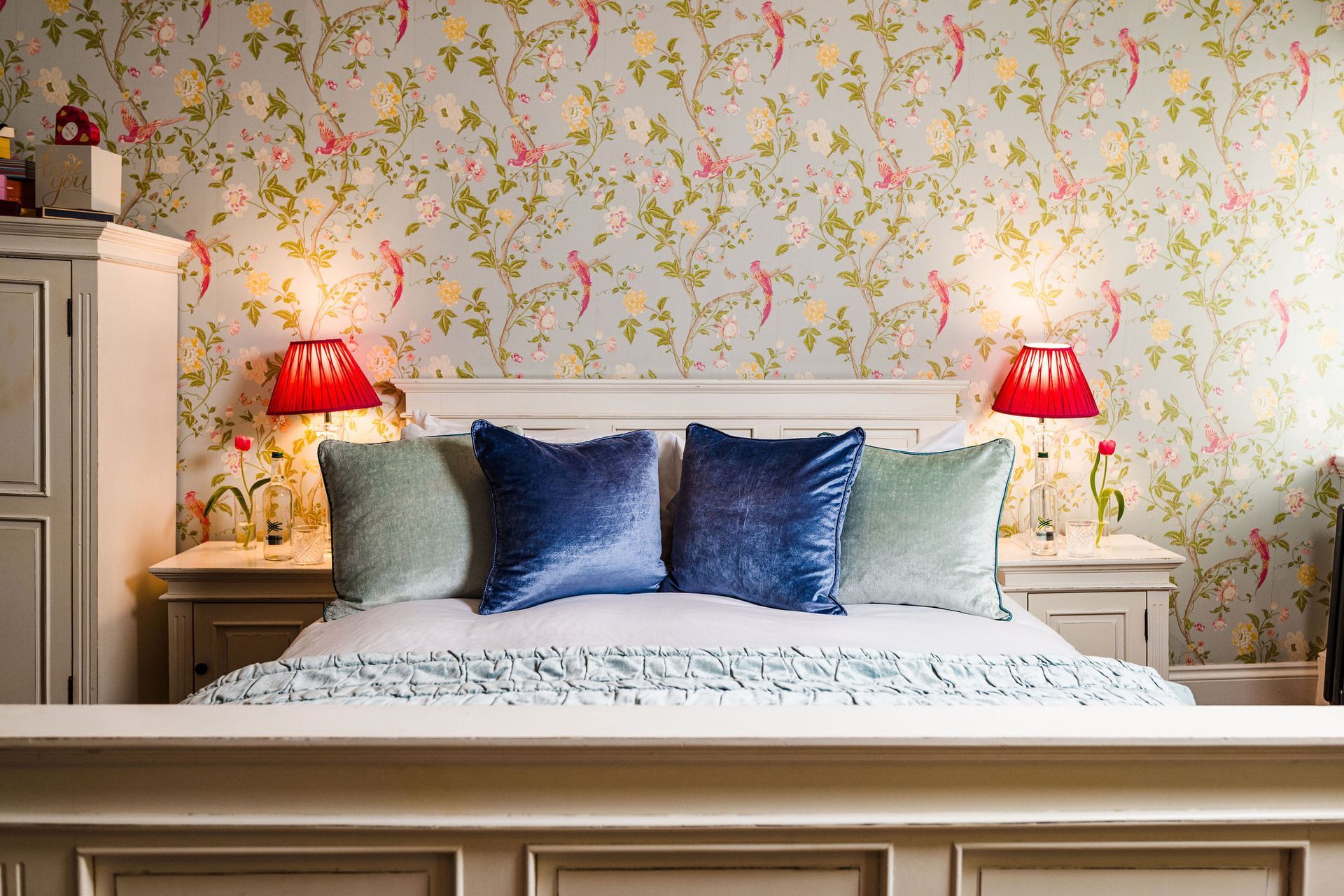 Cozy bed with blue pillows, red lamps, and floral wallpaper in a bright bedroom
