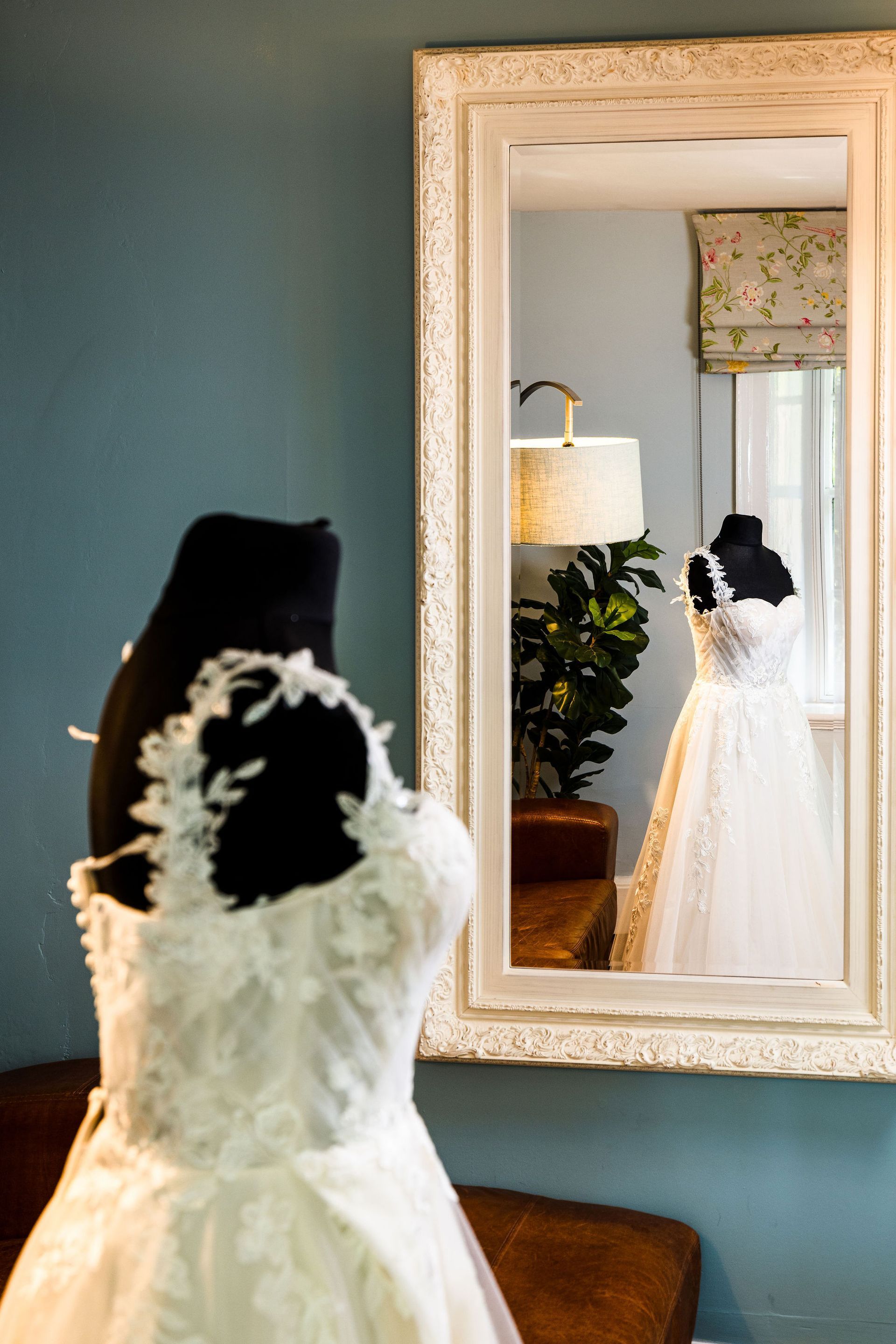 Wedding dress displayed in a mirror, with white flowers on the bodice and a black mannequin in a teal room