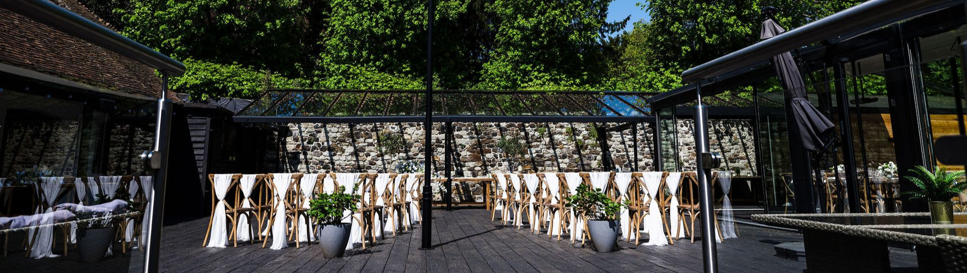 Rows of people in white robes standing in a shaded outdoor courtyard with stone walls and greenery
