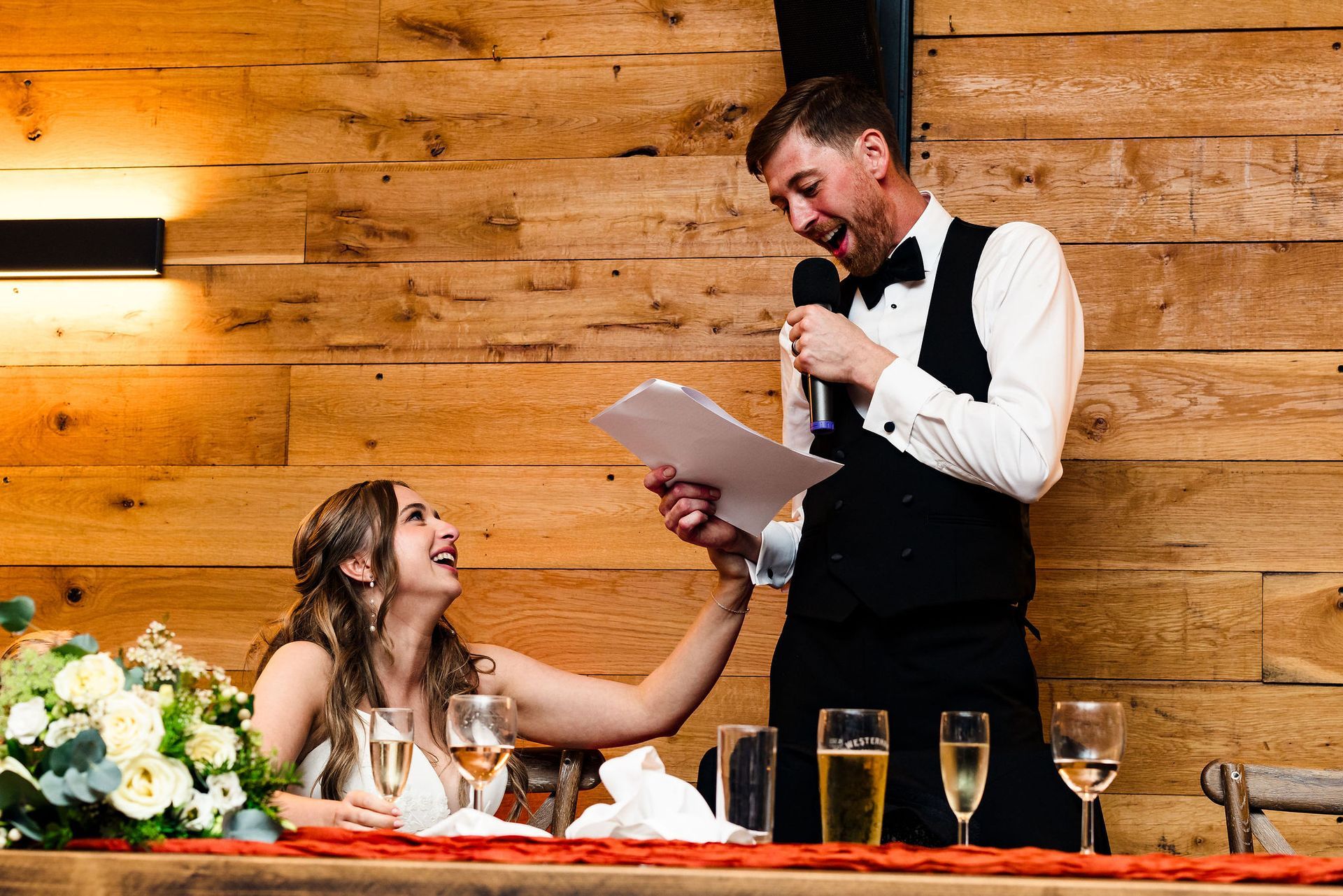 Man giving a toast with a microphone at a wedding reception while seated bride smiles and reaches up to him