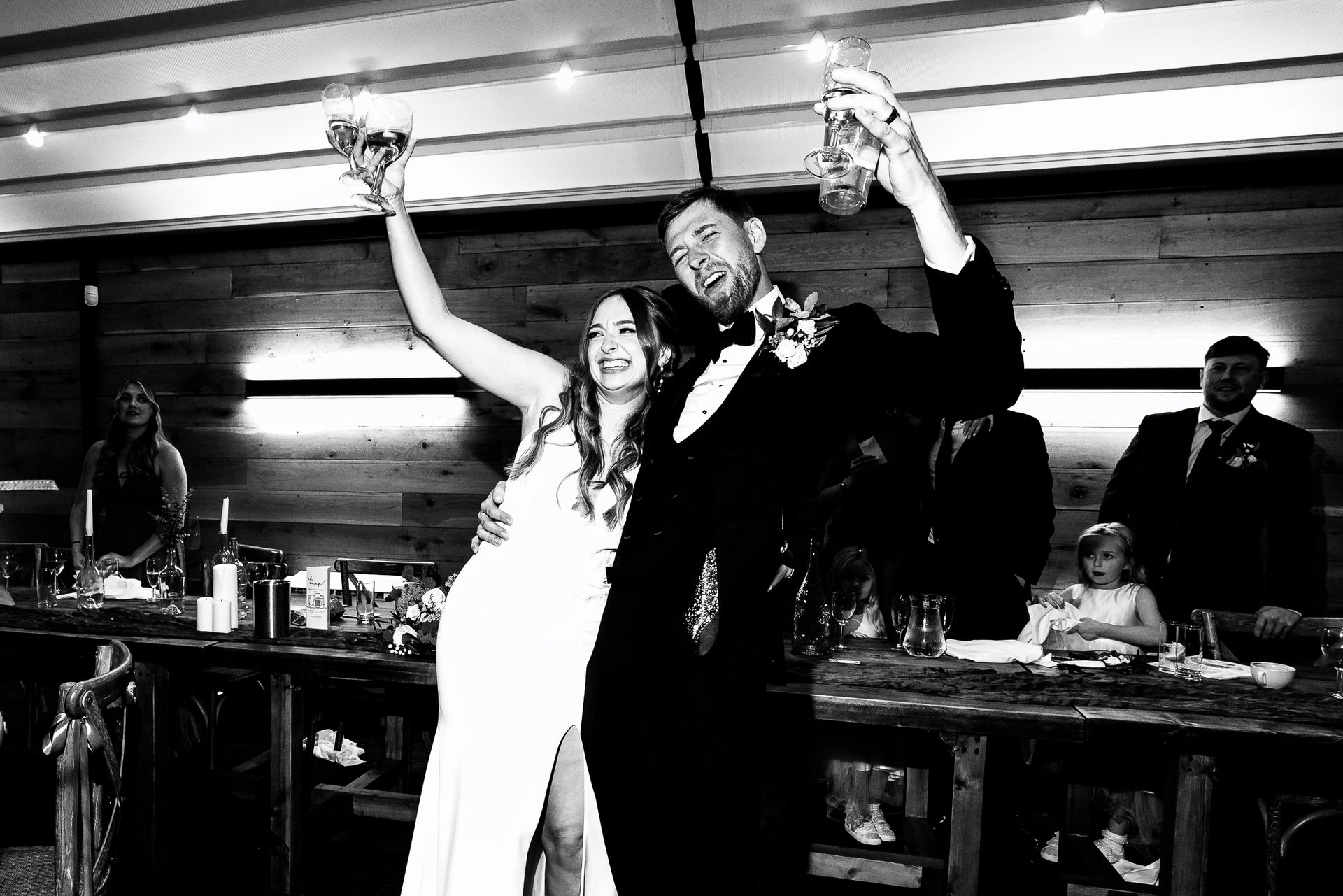 Smiling couple in formal wear raise glasses at a reception table, with guests and bottles in the background.