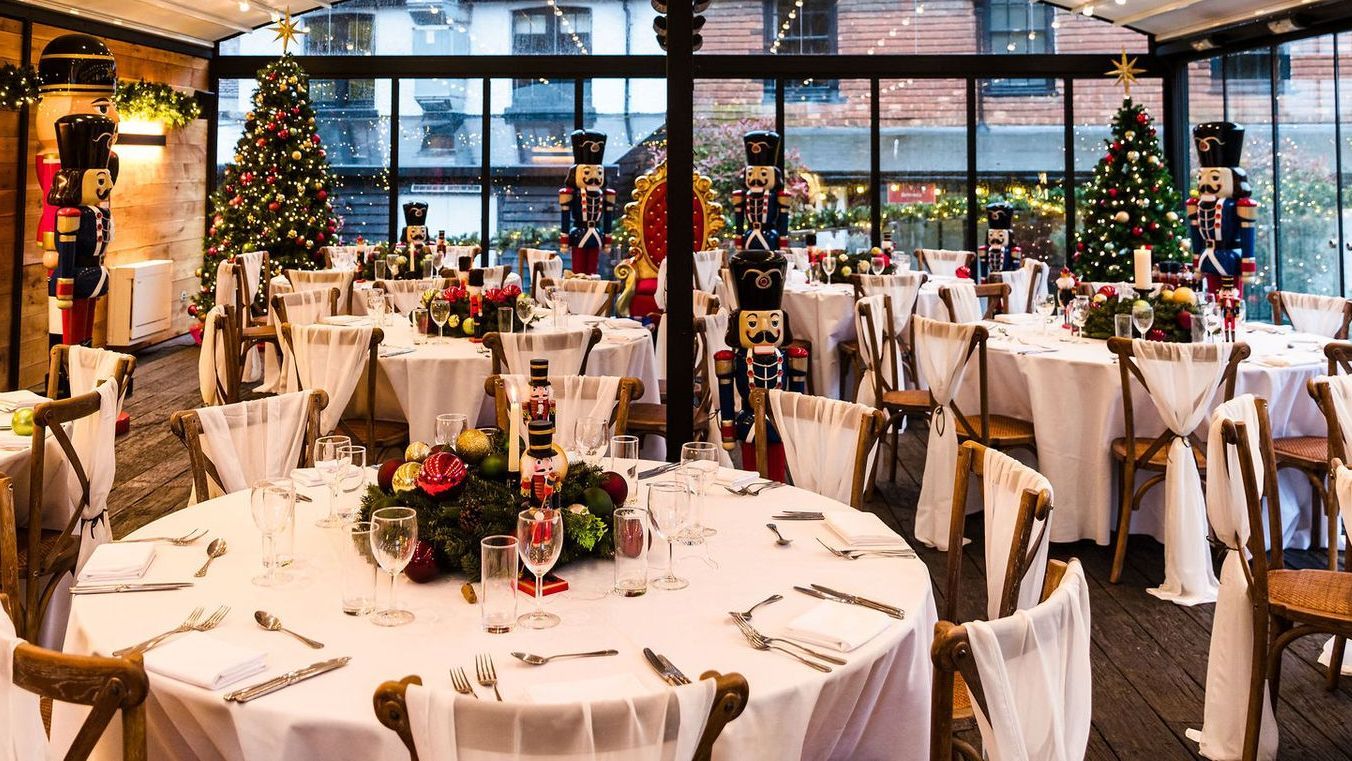 Festively decorated banquet hall with round tables, white chairs, and Christmas trees near large windows