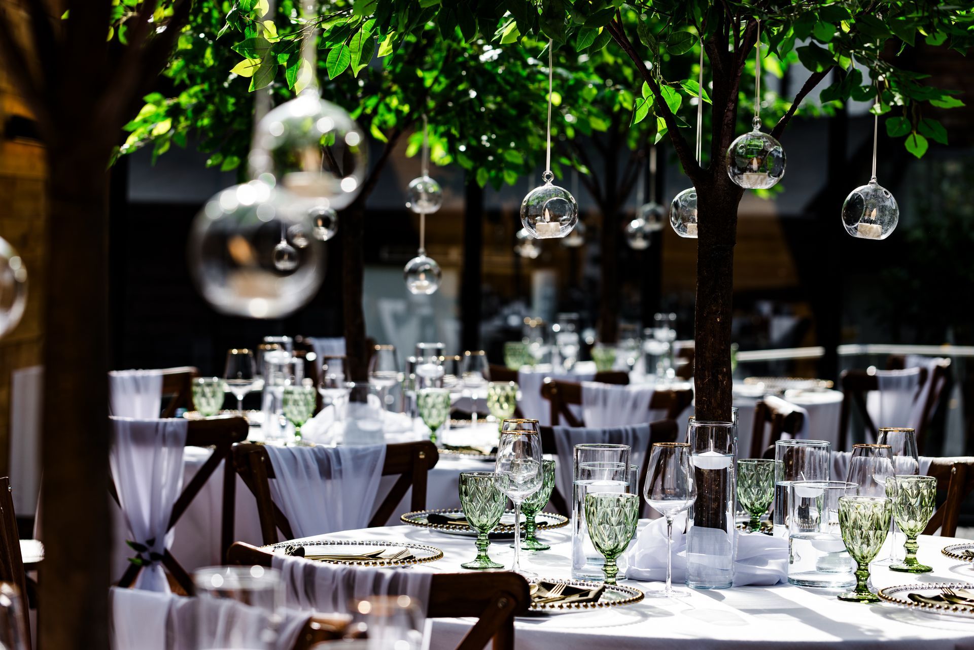 Outdoor dining tables under green trees with hanging glass lights and white tablecloths