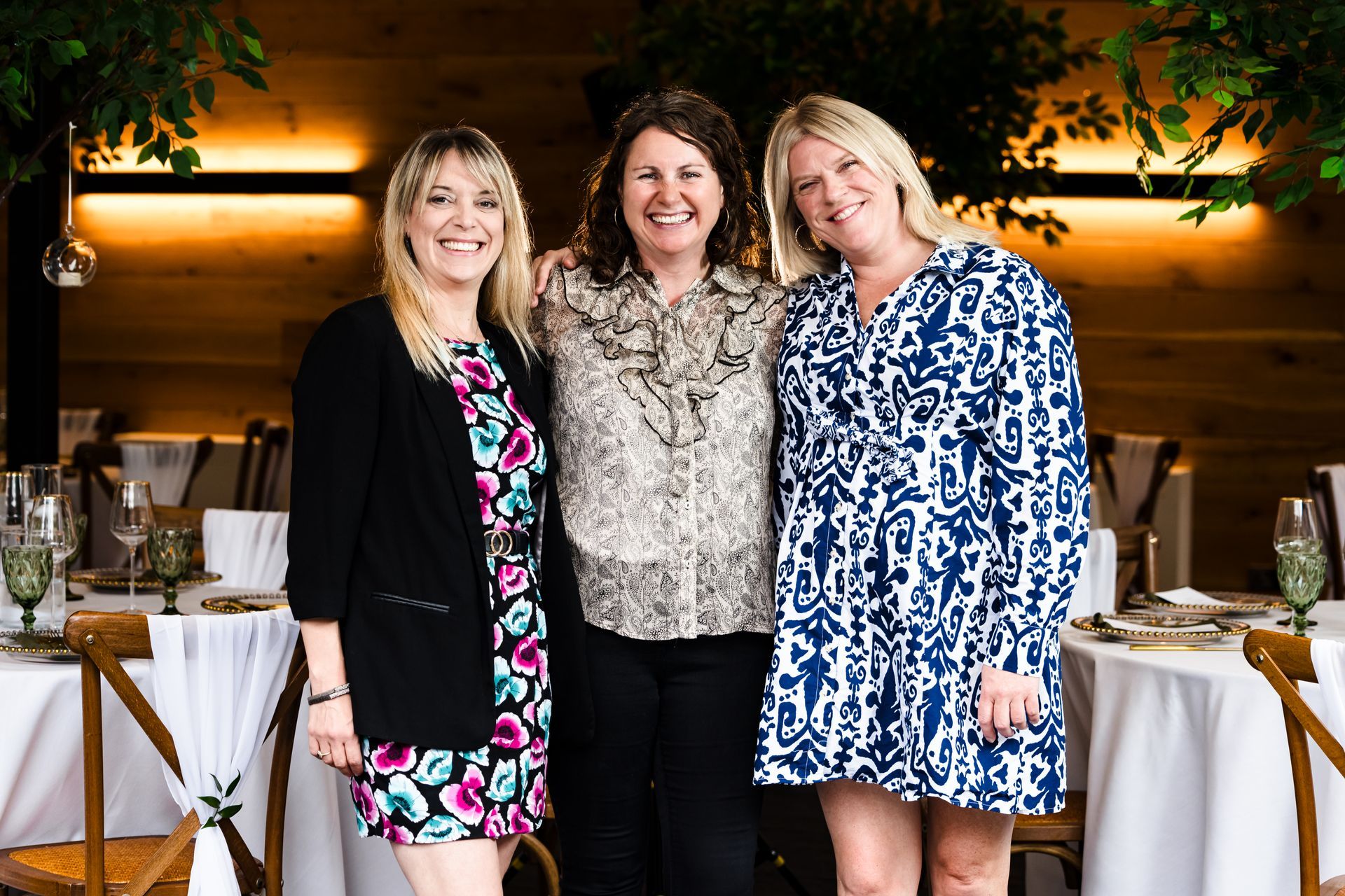 Three women posing together in a warmly lit restaurant with table settings and greenery backdrop