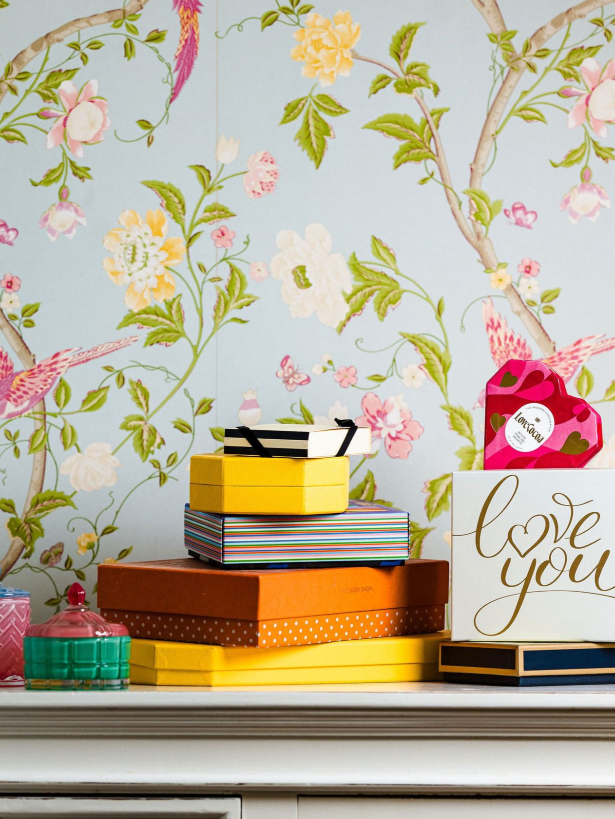 Decorative stack of colorful gift boxes with floral wallpaper and a “love you” sign on a shelf