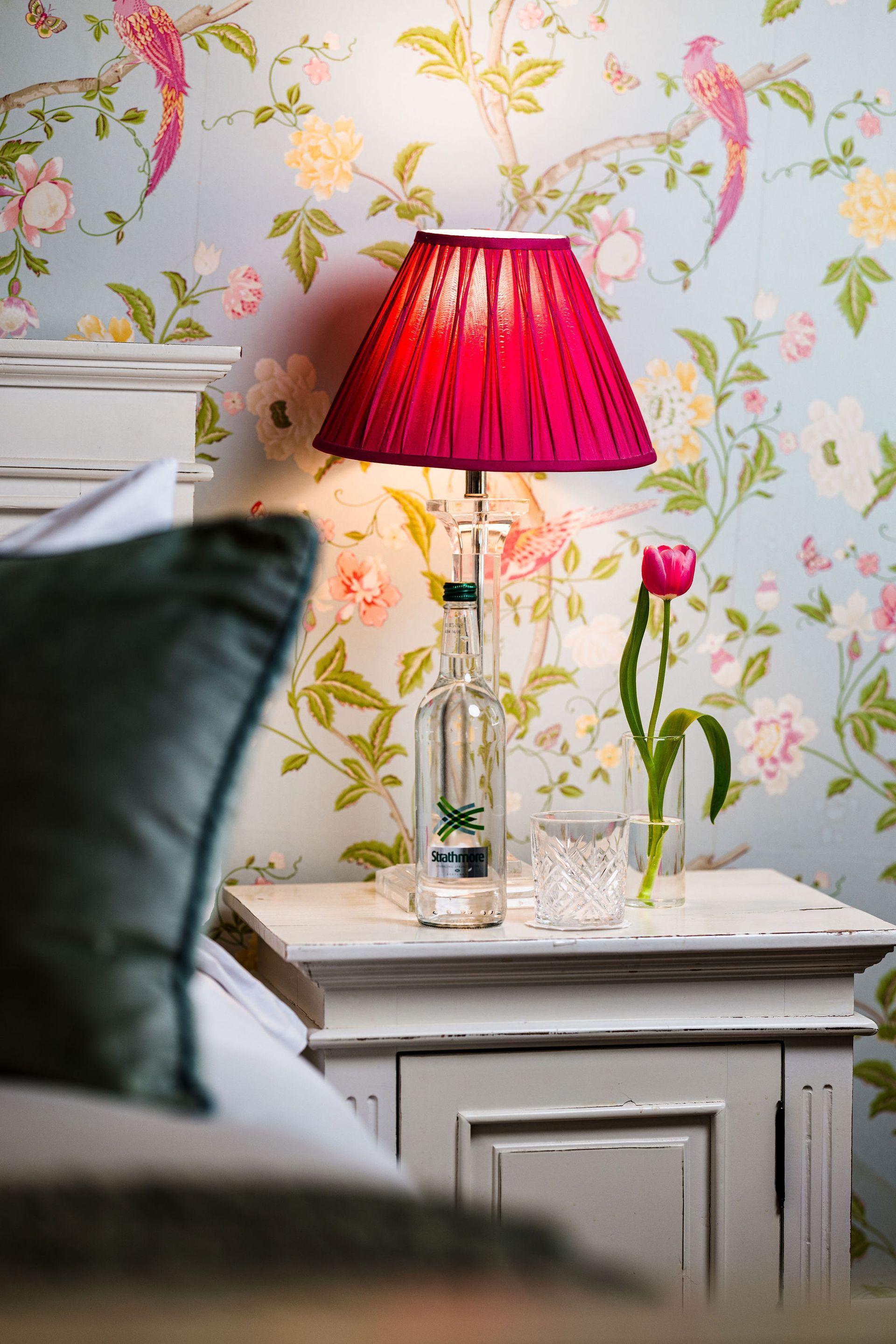 Pink lamp and tulip vase on a bedside table against floral wallpaper, with a bed in the foreground