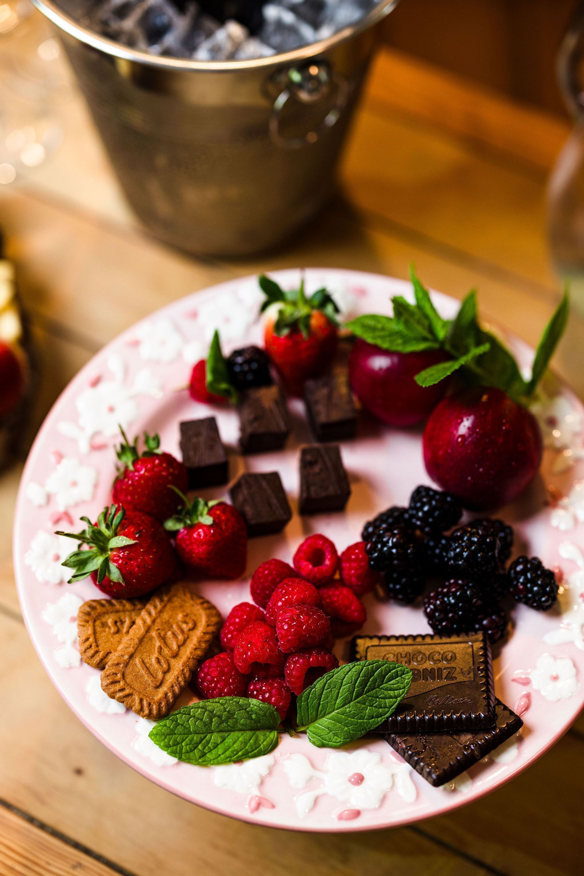 Plate of assorted chocolates and berries on a white floral plate, with mint leaves and a metal bucket in the background