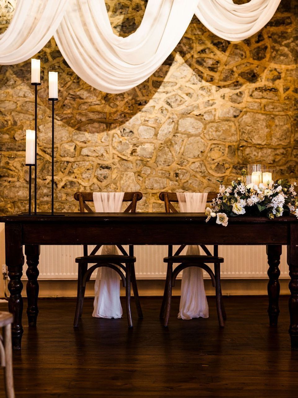 Elegant wedding reception table with draped white fabric, candles, and floral decor against a stone wall