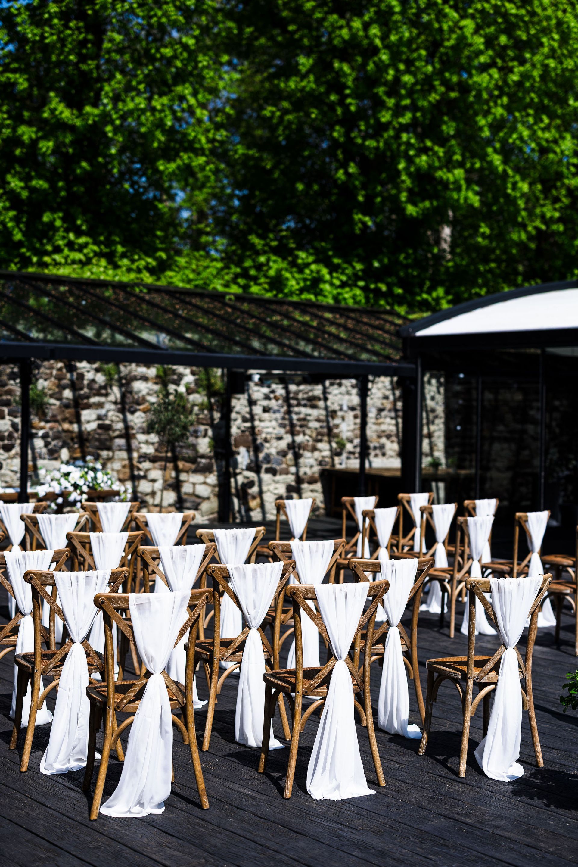 Outdoor event seating with gold chairs and white sashes on a patio beside a stone wall and greenery