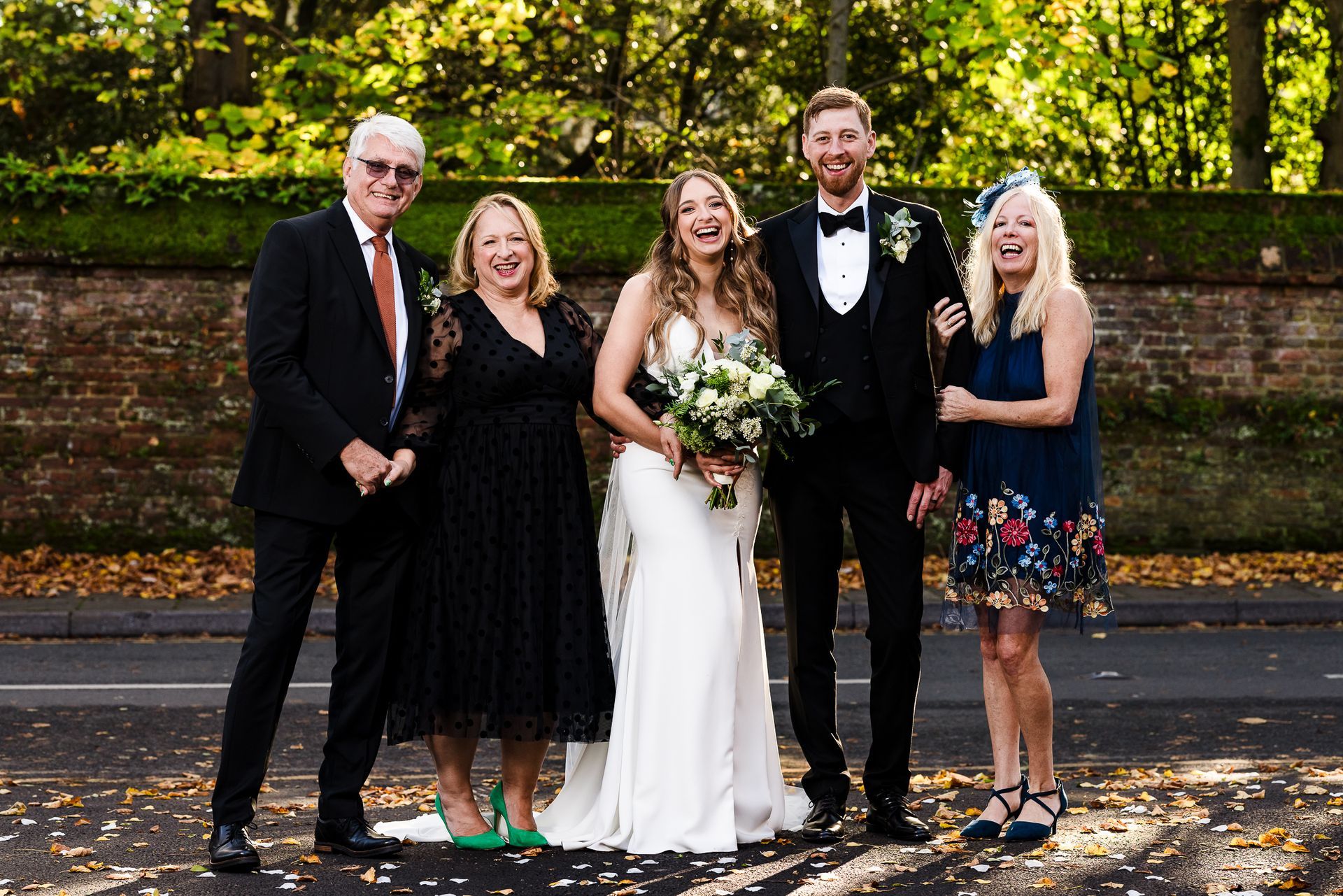 Wedding party standing outdoors with bride and groom centered, holding bouquet, on a leafy path.