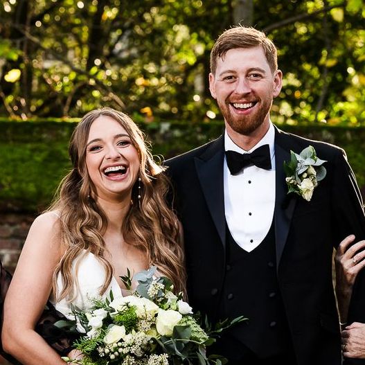 Smiling couple in wedding attire outdoors, holding a bouquet with greenery behind them