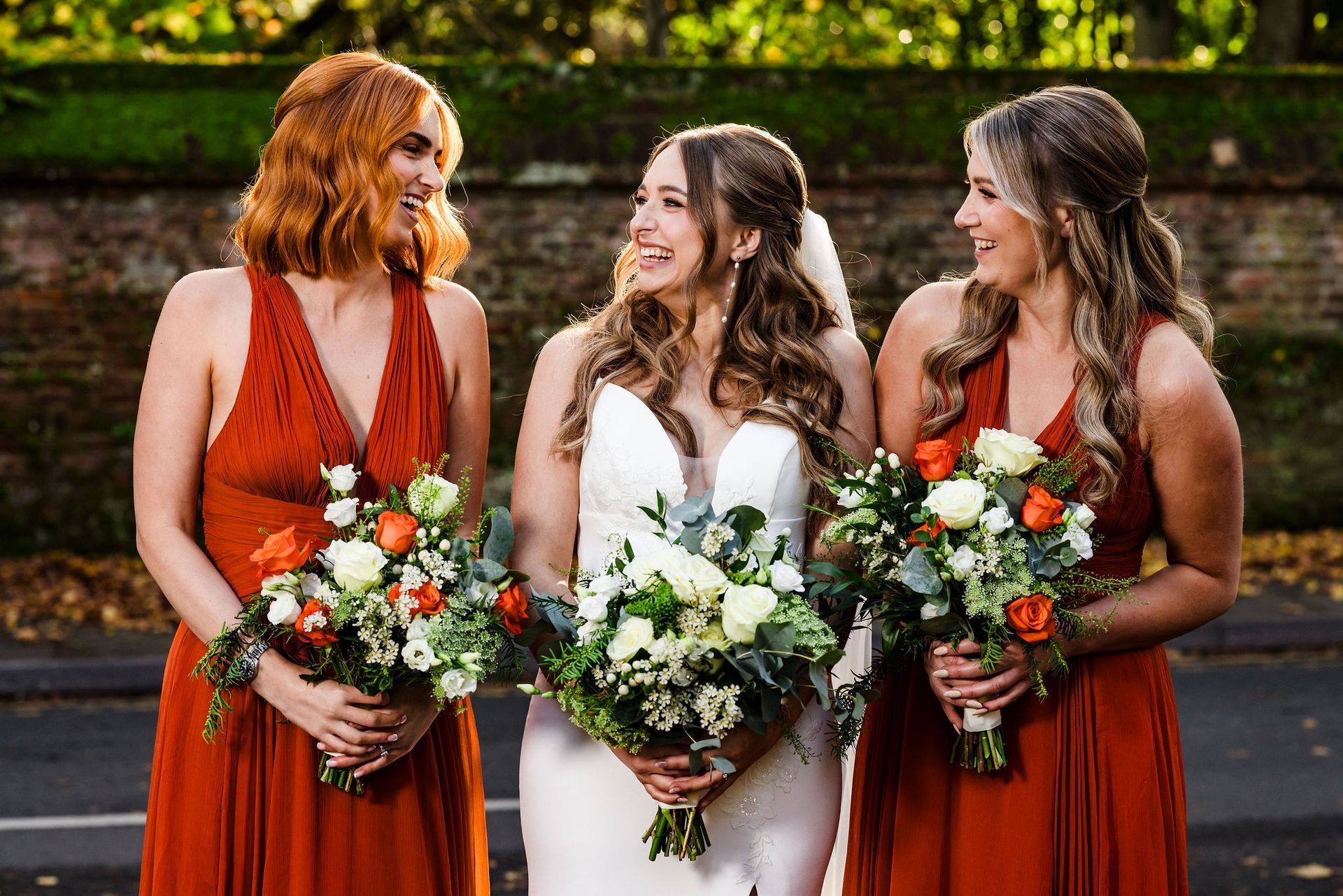 Three women in formal dresses holding bouquets, standing outdoors and smiling at each other.