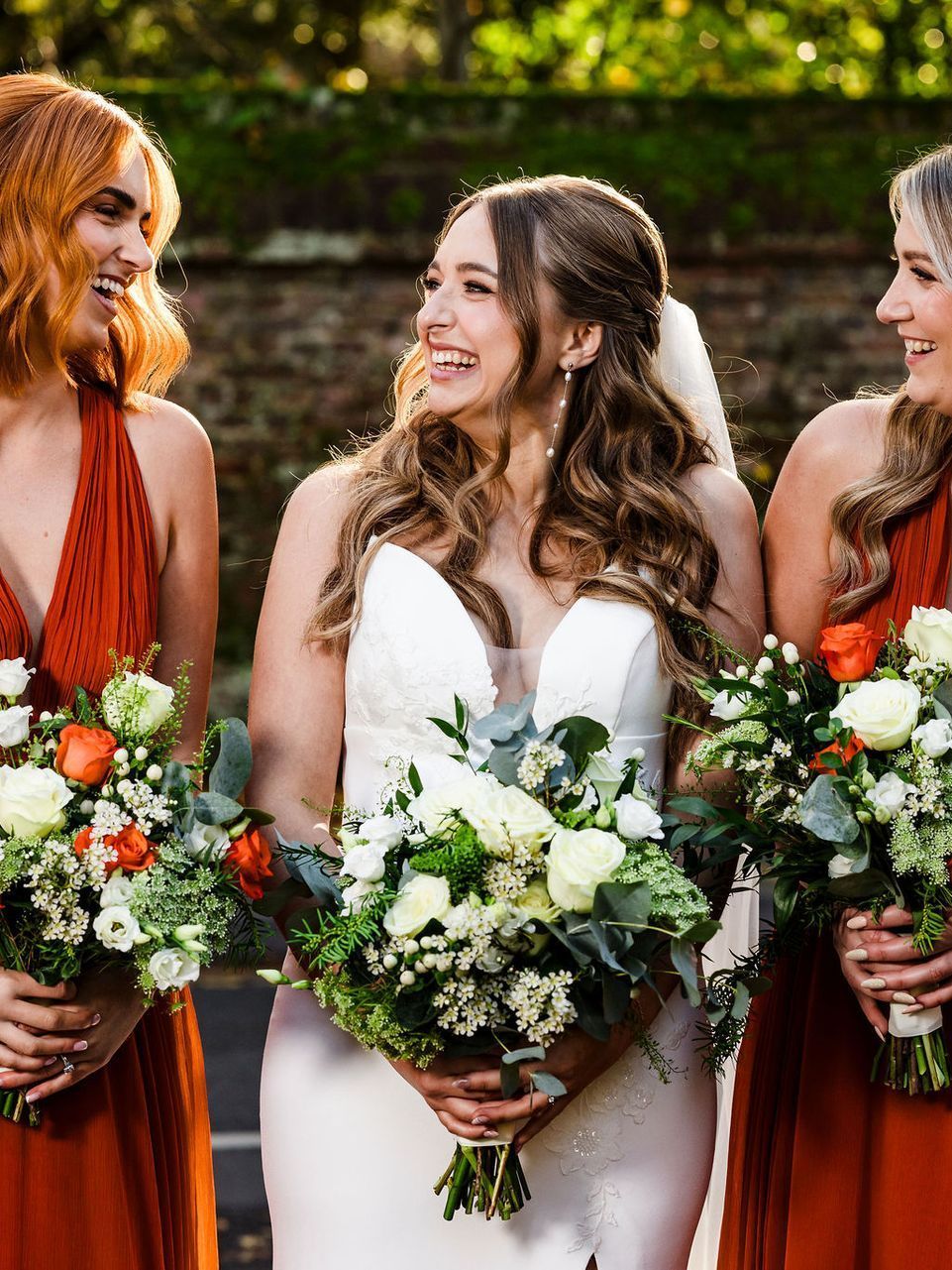 Three women in dresses holding flower bouquets, smiling outdoors at a wedding.