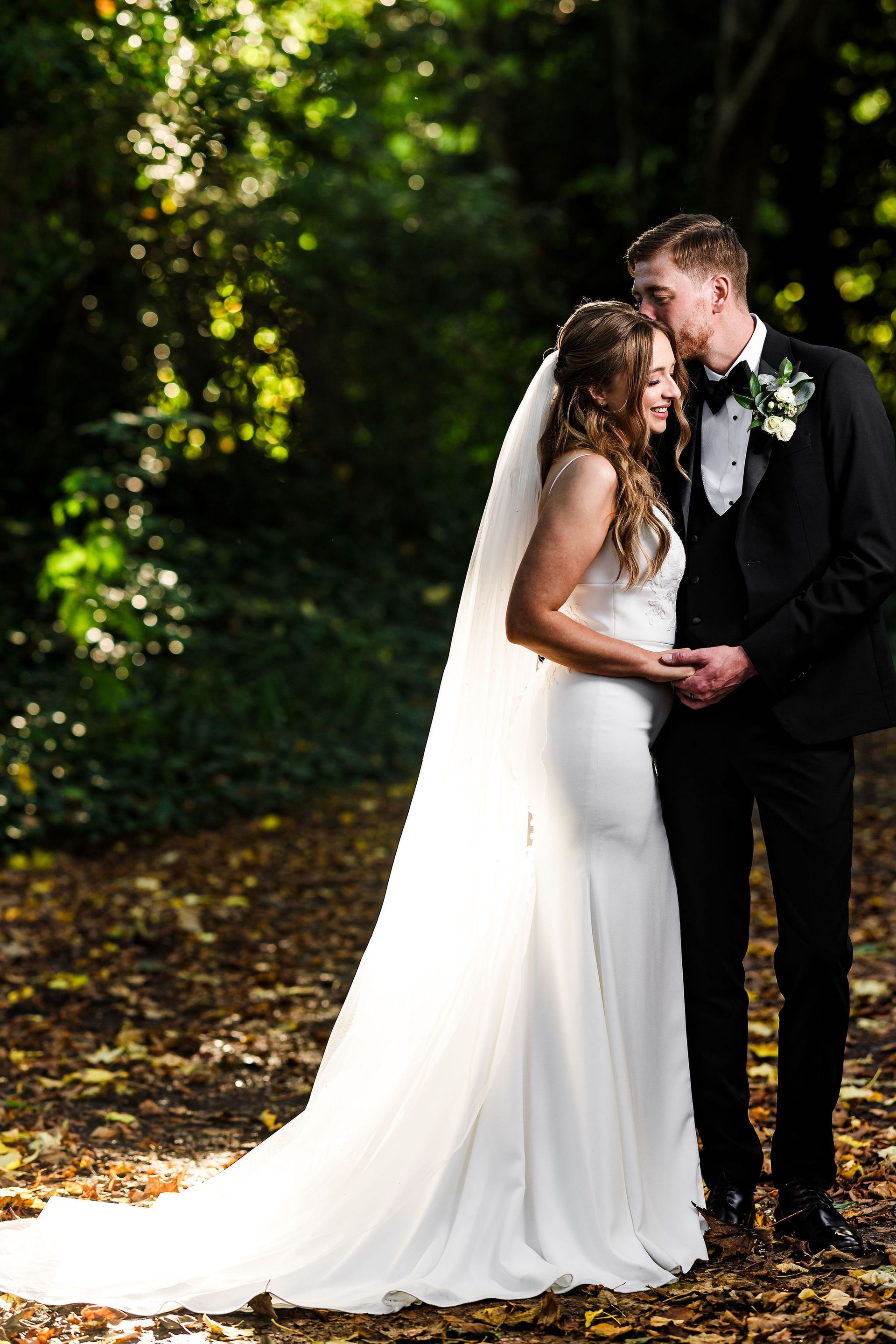Bride and groom embracing in a sunlit forest, with the bride’s long white dress flowing behind her.