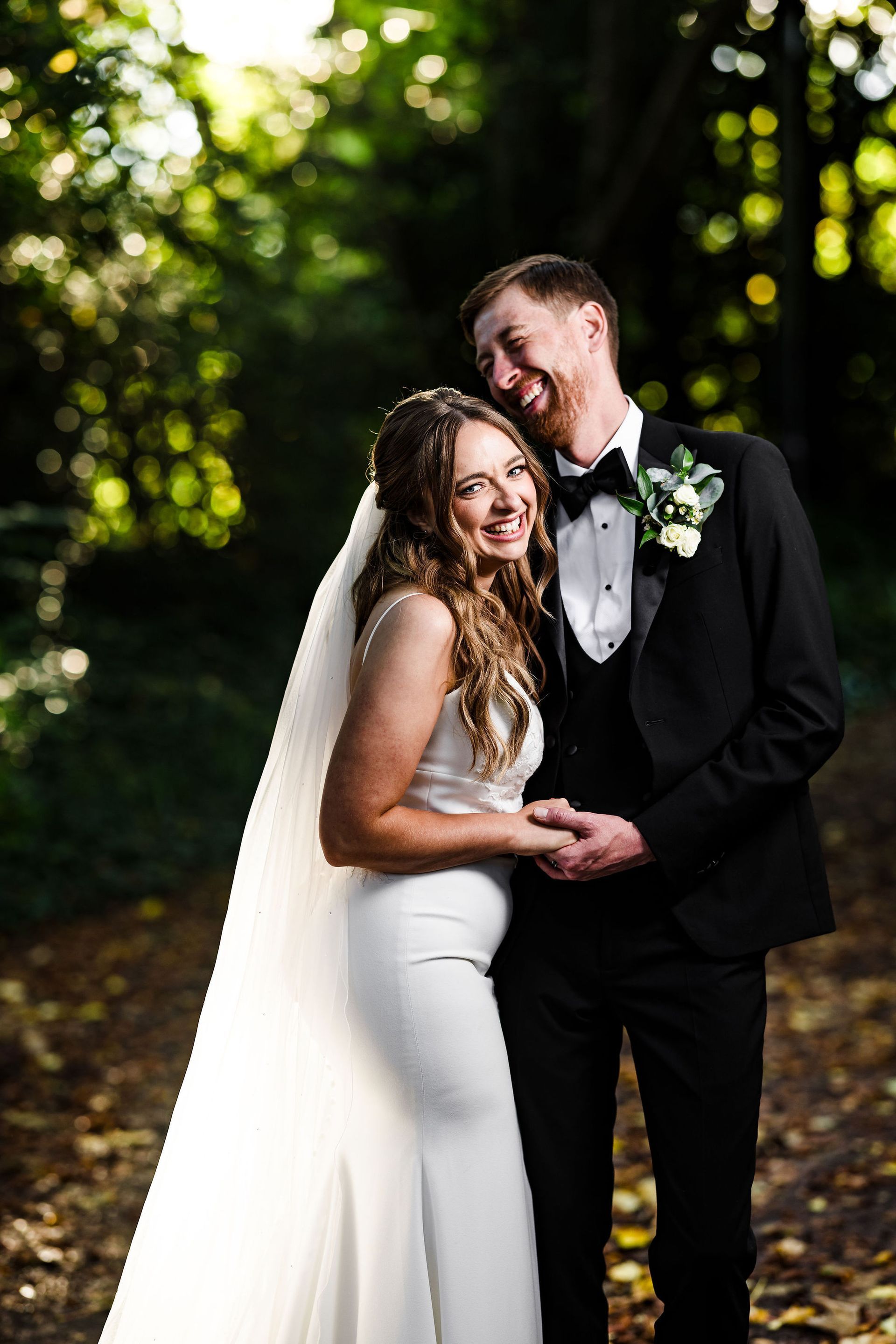 Newlyweds holding hands outdoors, smiling in wedding attire with the bride’s veil flowing behind her.
