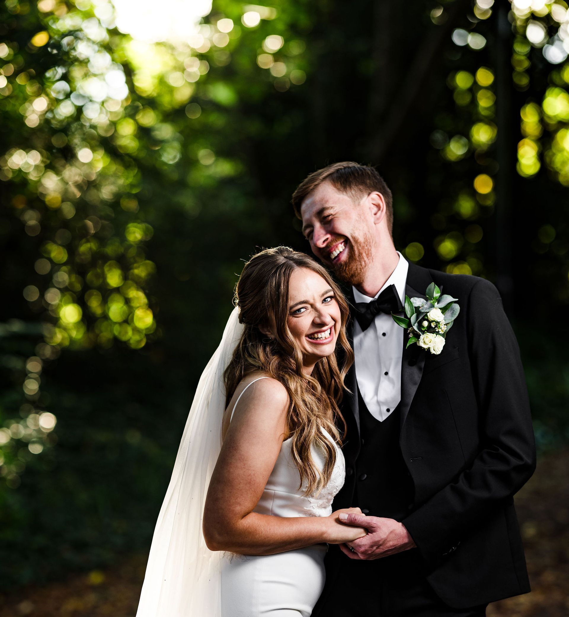 Bride and groom smiling in a dark outdoor garden, holding hands under soft natural light