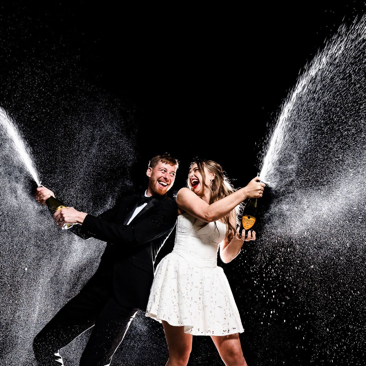 Two people in formal clothes spraying champagne against a black background