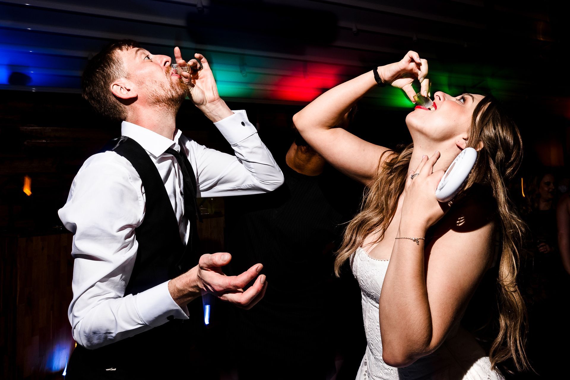 Two people dancing and drinking at a dimly lit party, with colorful lights overhead.