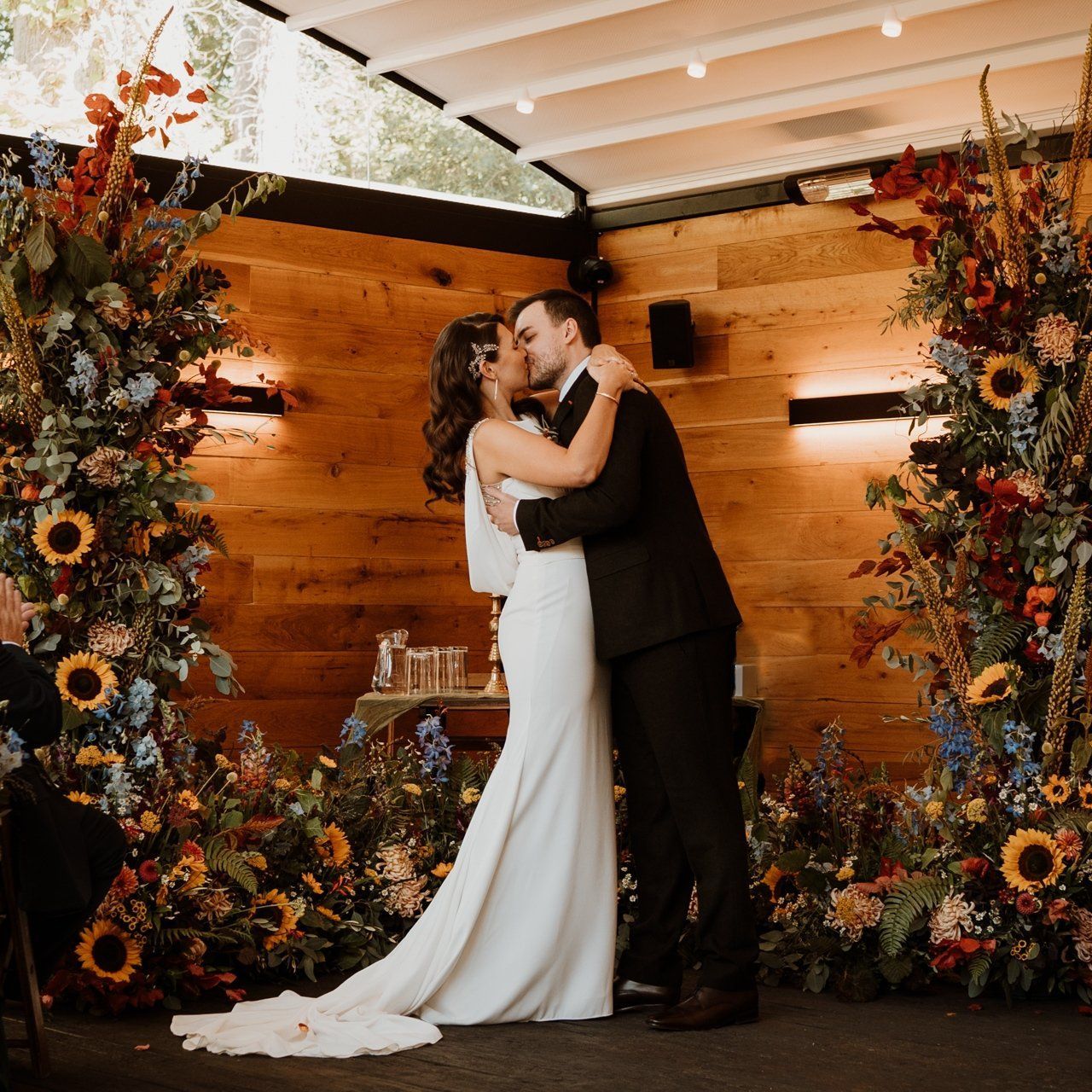 Couple kissing at a rustic wedding ceremony with floral arrangements and warm wood backdrop