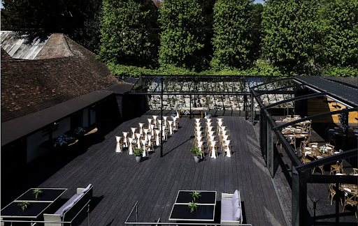 Outdoor rooftop dining deck with rows of tables and white chairs beside a black building and trees