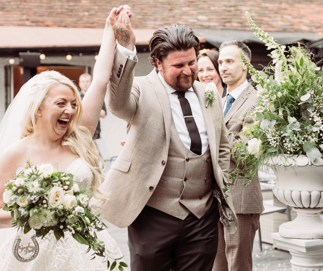 Joyful couple in beige wedding attire raising hands outdoors at a garden wedding, holding bouquets