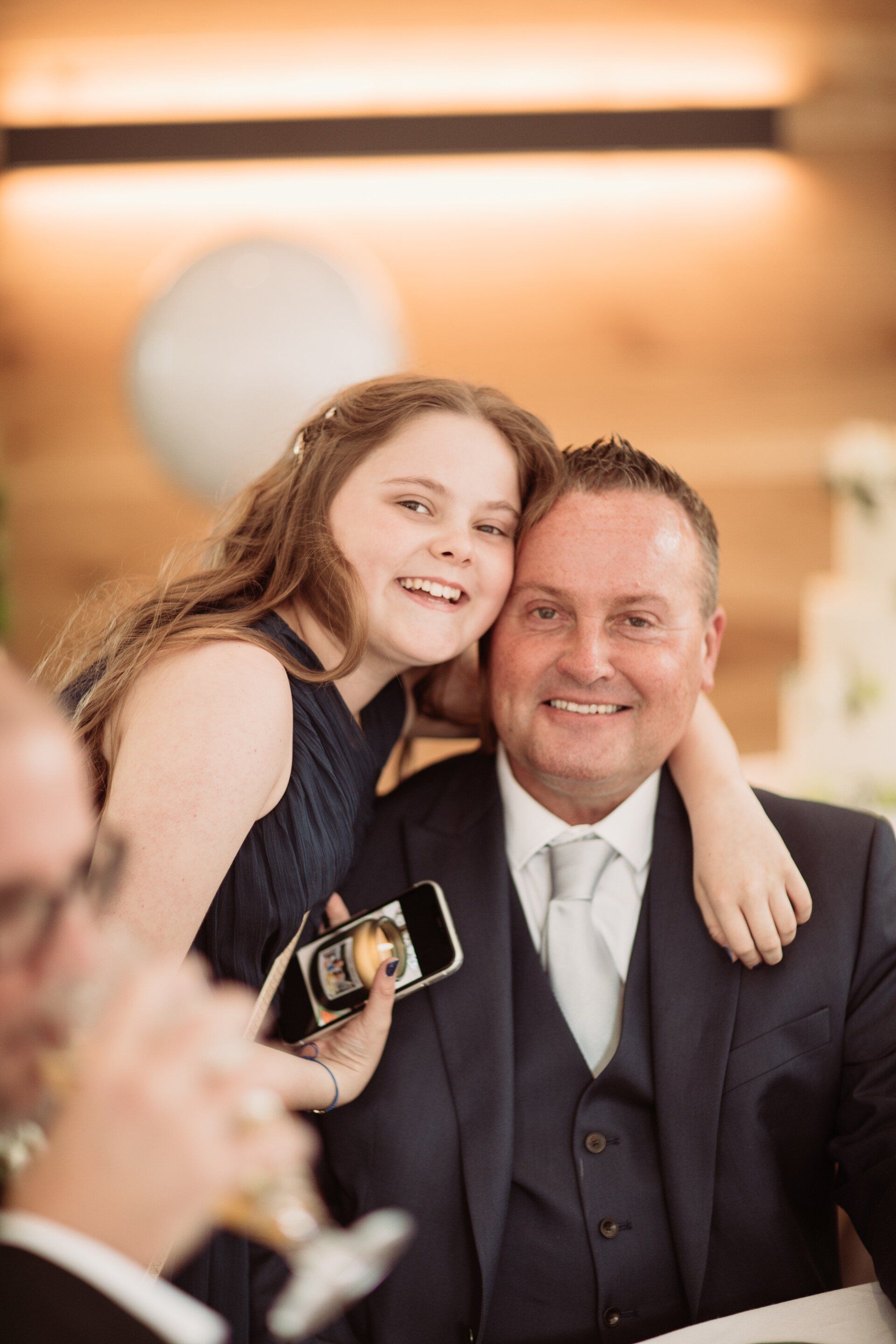 Smiling girl hugging a man in a dark suit at a formal event, with a phone visible in the foreground