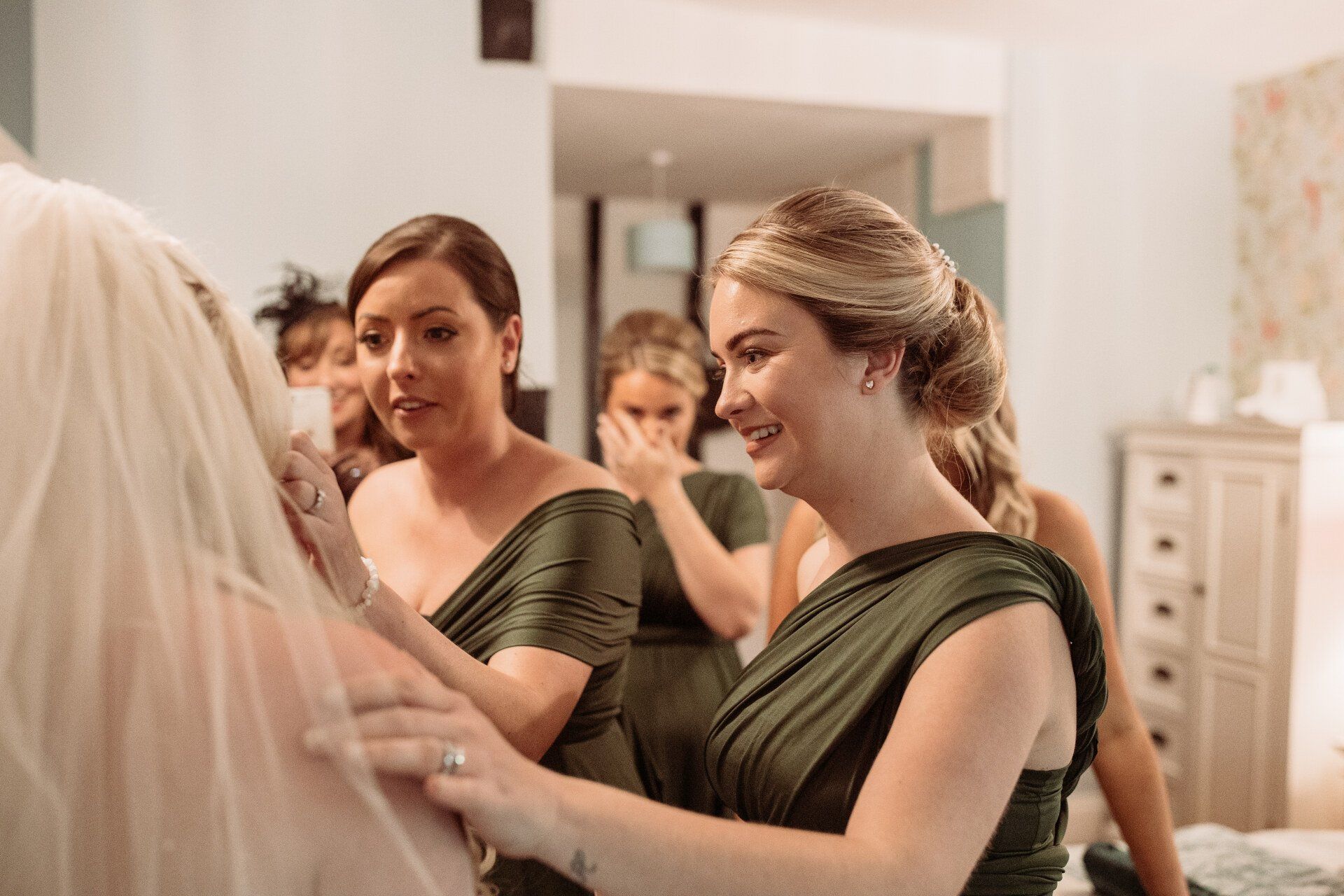Bridesmaids in green dresses help a bride with her veil in a bright dressing room