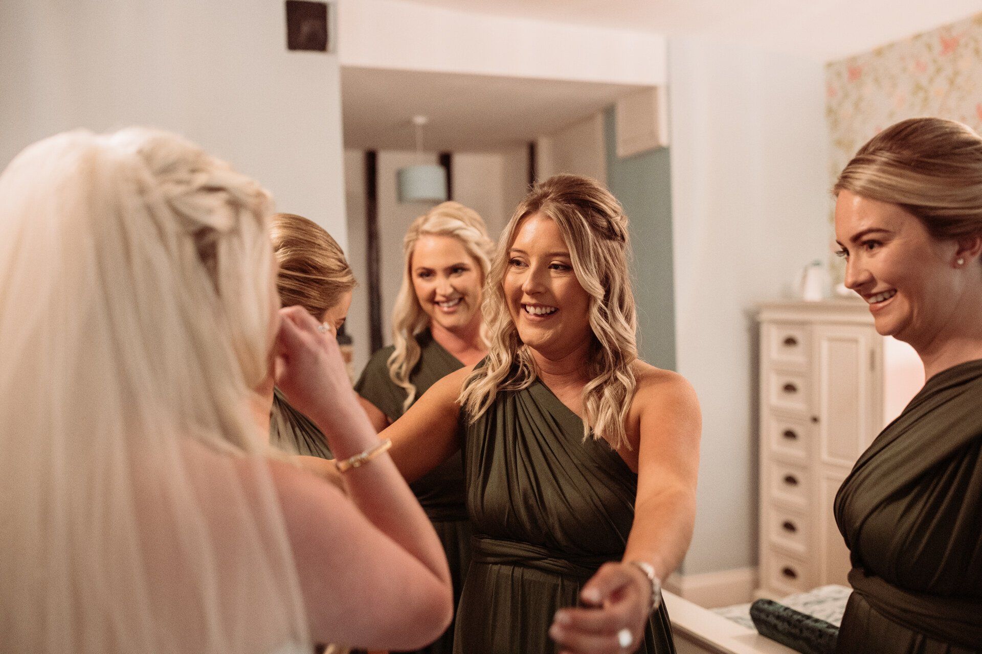 Five women smiling and chatting in a dressing room, with one in a dark green dress in the center