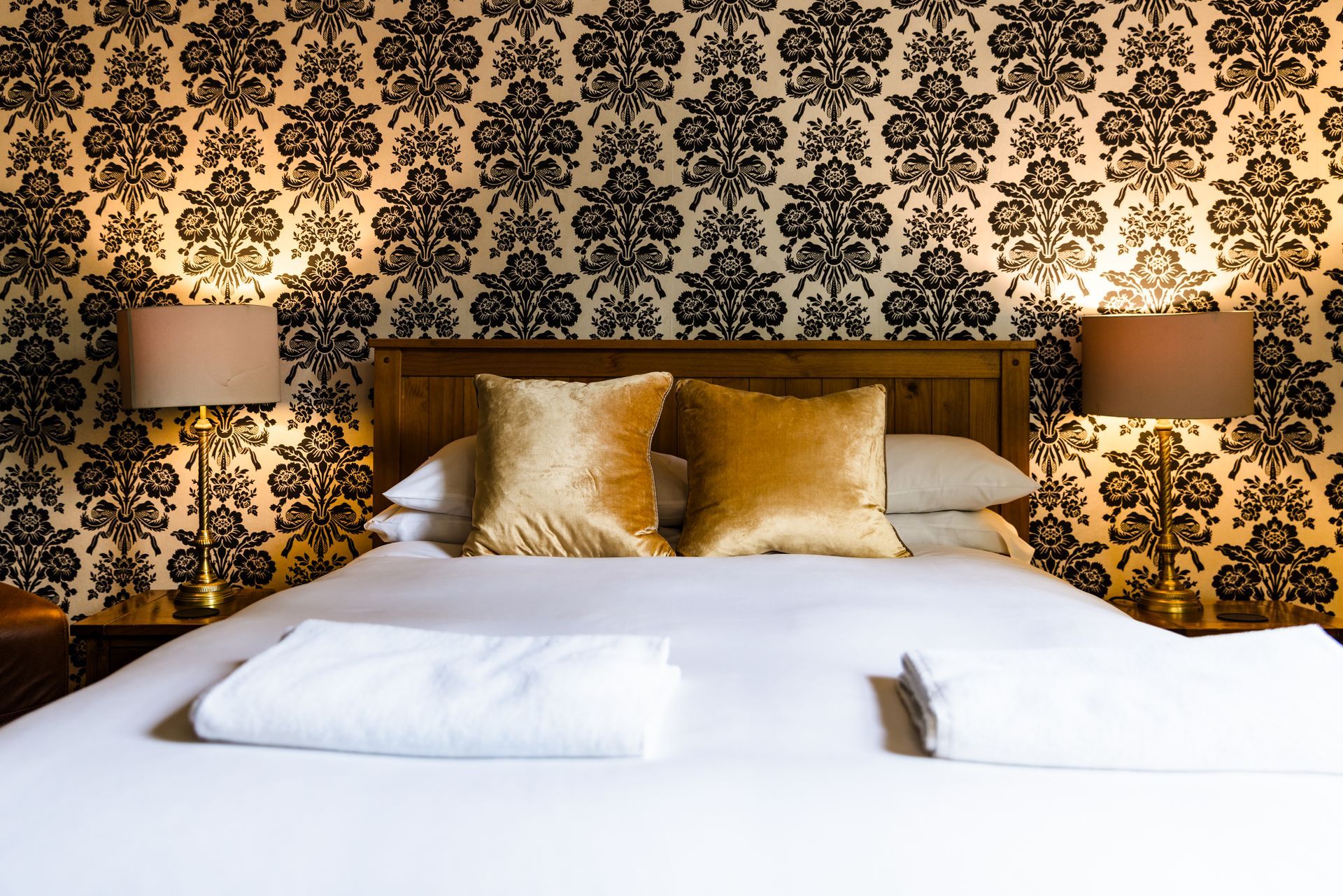 King-size bed with white linens, gold pillows, and bedside lamps against ornate black-and-gold wallpaper