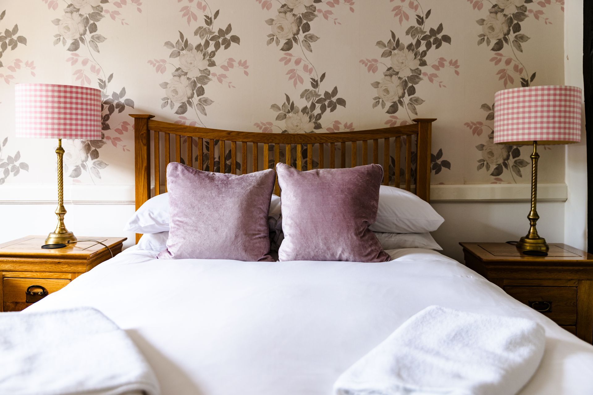 Cozy bed with floral wallpaper, pink lamps, and lavender pillows in a softly lit bedroom