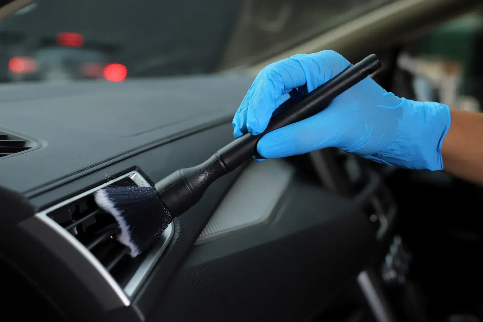 Hand in blue glove uses a brush to clean a car's dashboard vent.