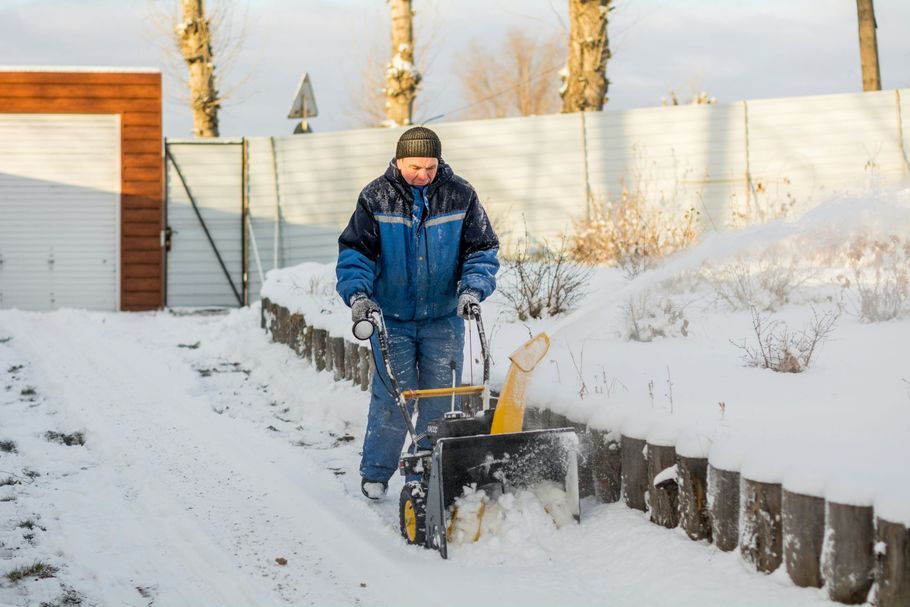 Person using a snowblower on a snowy driveway beside a white fence and house