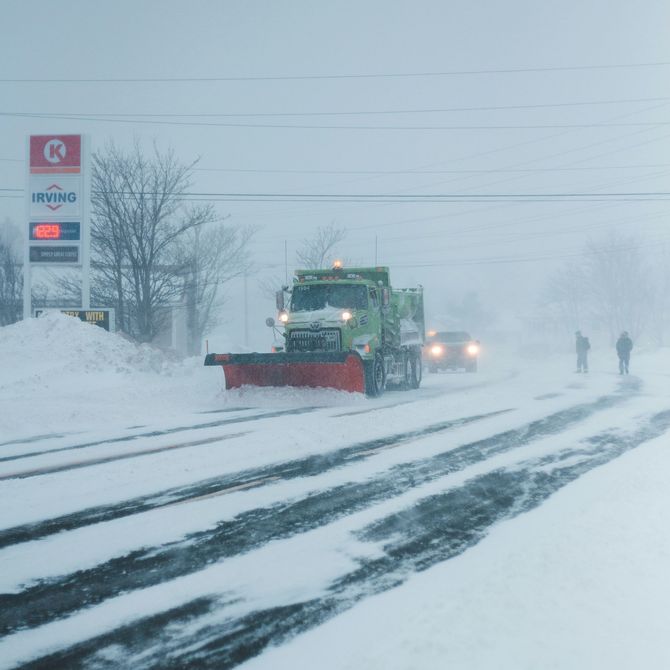Snowplow clearing a snowy road in a blizzard, with headlights and two pedestrians in the distance