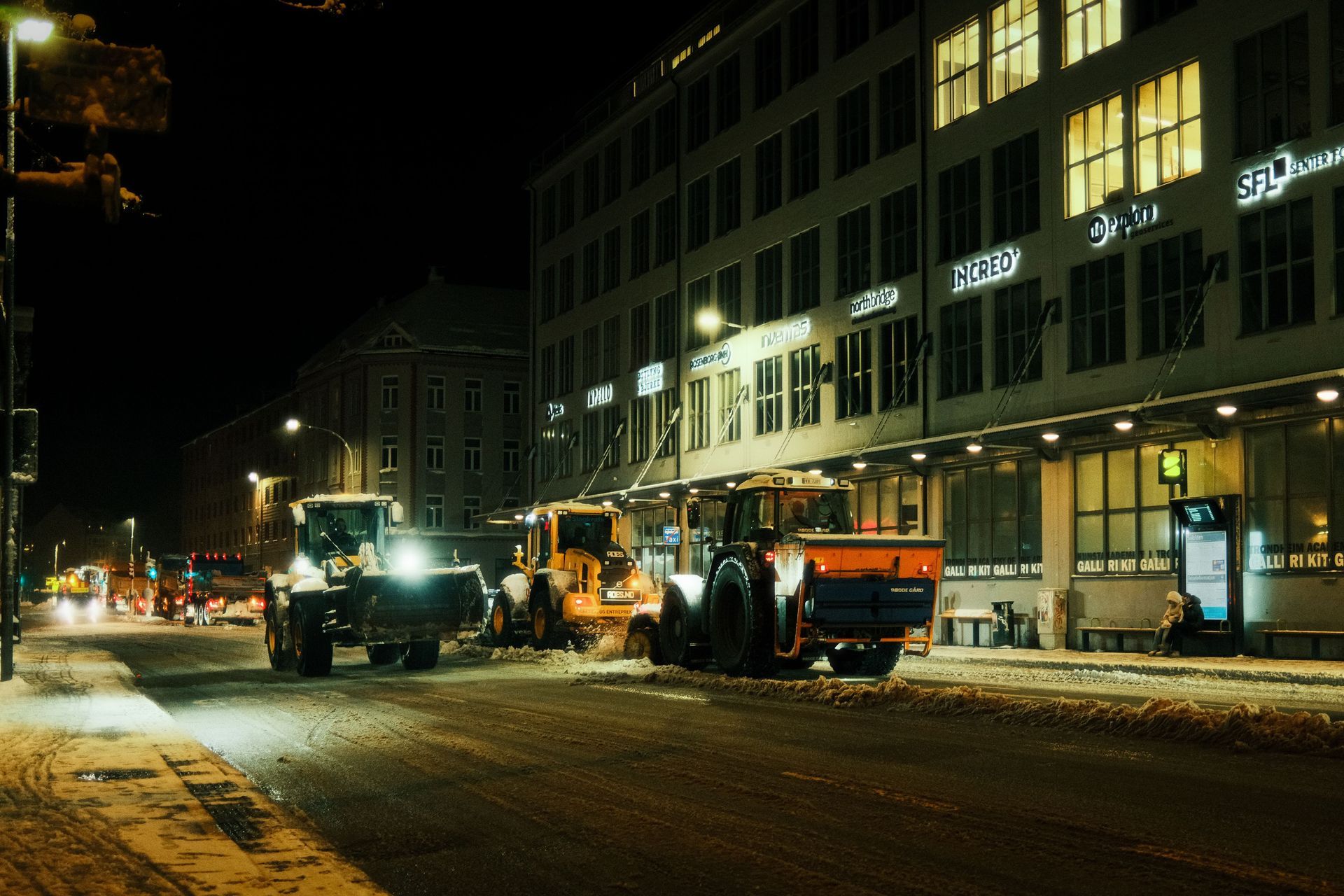 Snowplows and construction vehicles clearing a snowy city street at night beside lit buildings.