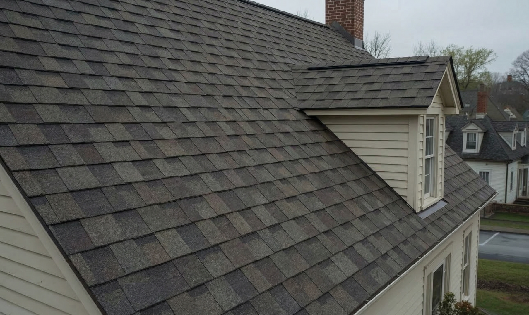 A roof with textured gray shingles and a beige dormer window against an overcast sky.